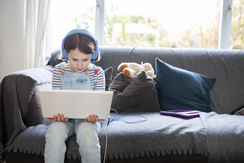 Kind mit Kopfhörer und Laptop auf einem Sofa sitzend.
